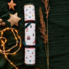 Three stacked cylindrical gift wraps with festive prints (polka dots and Christmas icons) tied with teal ribbons, set on dark green fabric beside star ornaments and warm fairy lights.