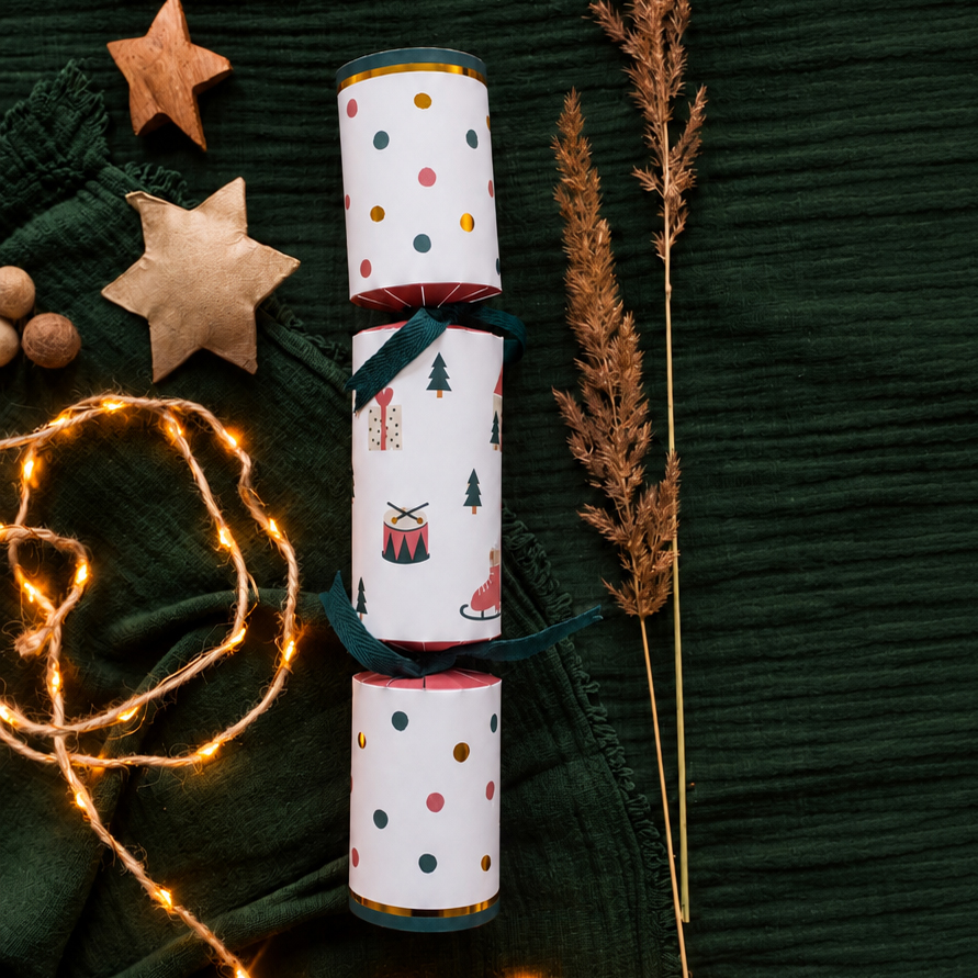 Three stacked cylindrical gift wraps with festive prints (polka dots and Christmas icons) tied with teal ribbons, set on dark green fabric beside star ornaments and warm fairy lights.