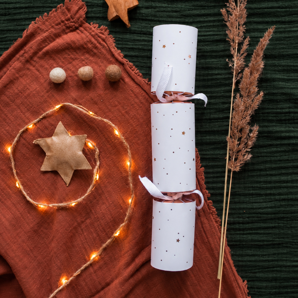 4145knallbonbonssterne Avayves Feine Papeterie Und Geschenke Für Babys Und Kids Flat lay of holiday crafts: white star-patterned rolled papers tied with pale ribbons, warm fairy lights, a felt star on orange fabric, and dried grasses on dark green backdrop.