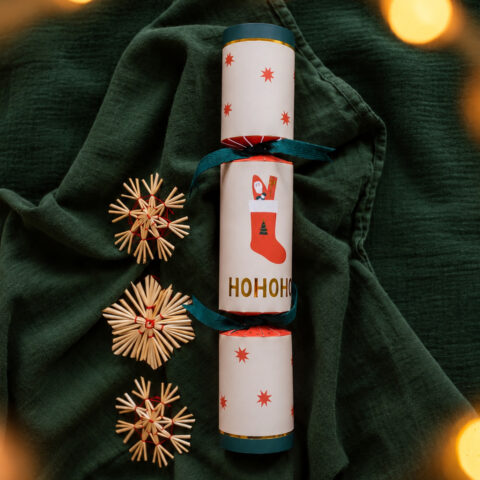 Vertical stack of three Christmas crackers on dark green fabric, with teal ribbons and red star patterns, surrounded by straw star ornaments and warm bokeh lights.