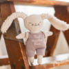 Soft beige plush bunny with long ears wearing a light purple romper that reads 'Mutmacher Hase', perched on a wooden chair.