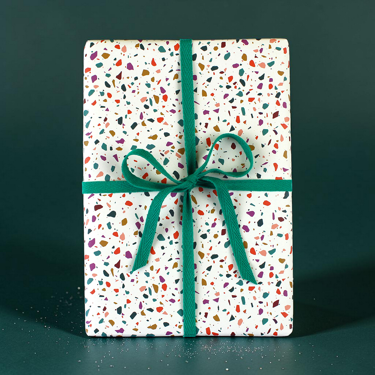 Gift box wrapped in white confetti-pattern paper with a teal ribbon and bow on a dark green background.