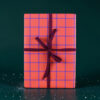 Wrapped gift box with pink-orange plaid pattern and purple ribbon against a dark green background, festive and ready to give.