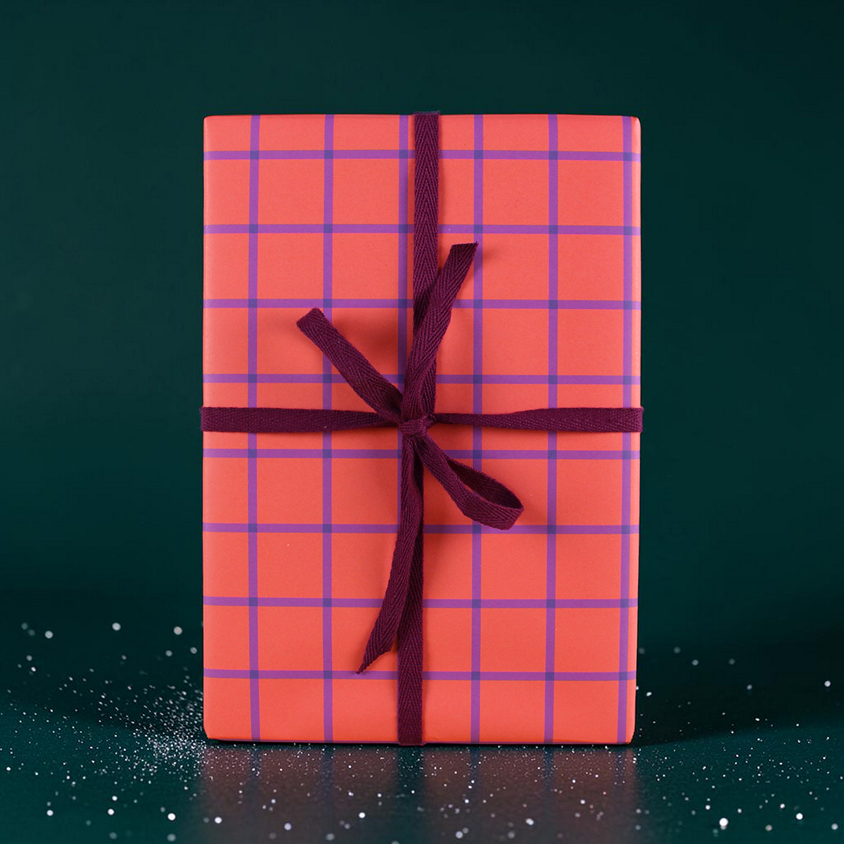 Wrapped gift box with pink-orange plaid pattern and purple ribbon against a dark green background, festive and ready to give.