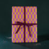 Rectangular gift box wrapped in purple and yellow checker paper with a burgundy ribbon against a dark green background.