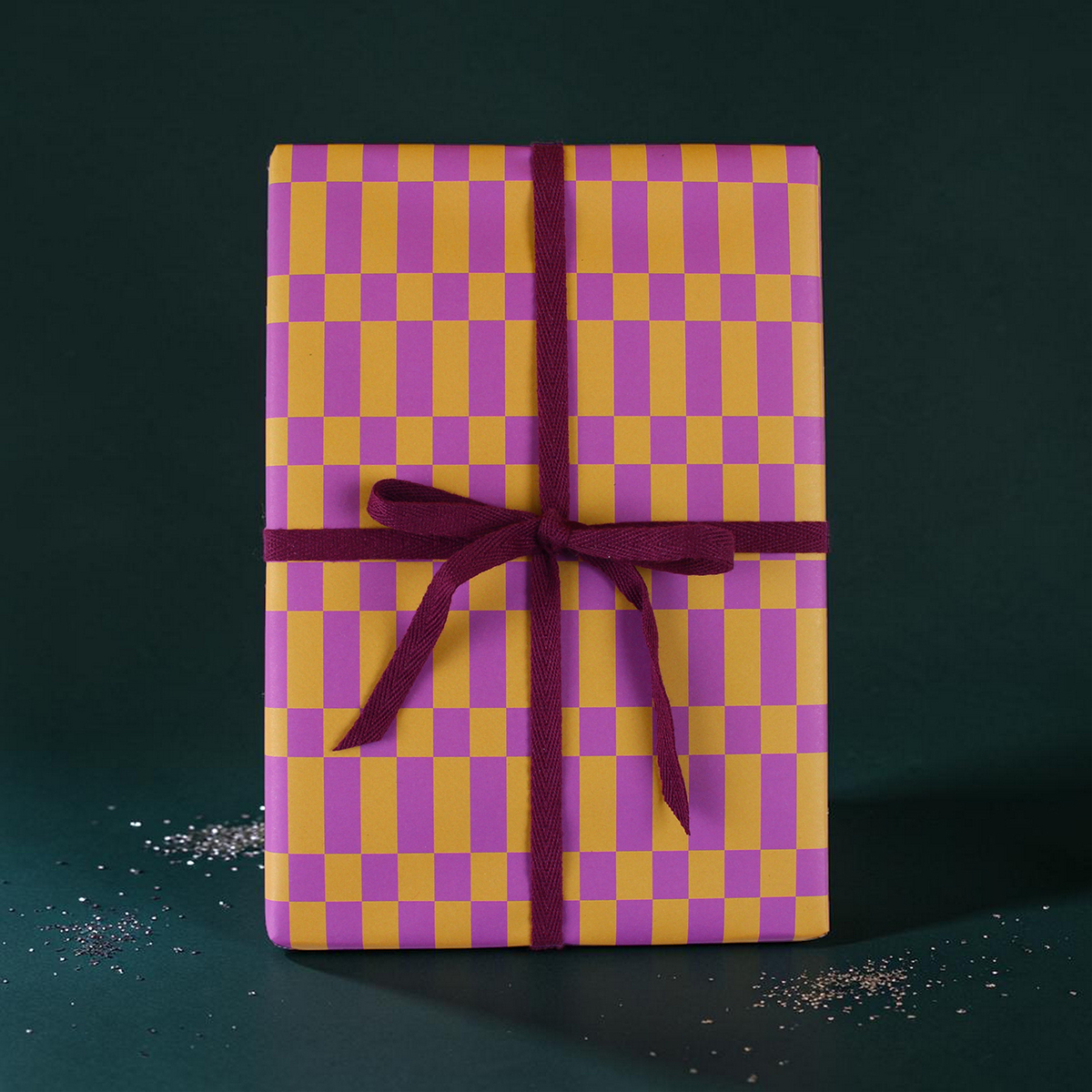 Rectangular gift box wrapped in purple and yellow checker paper with a burgundy ribbon against a dark green background.