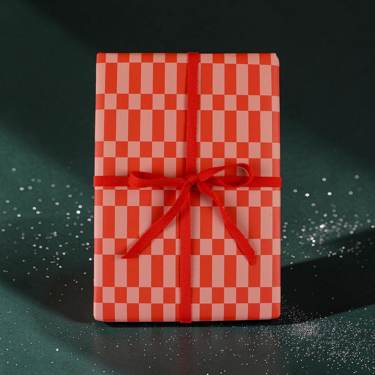 Gift box wrapped in red-and-pink checker-pattern paper with a red ribbon bow on a dark background for gifting.