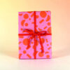 Pink gift box wrapped in patterned paper with a red ribbon and bow against a light yellow background.