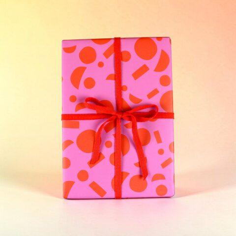 Pink gift box wrapped in patterned paper with a red ribbon and bow against a light yellow background.