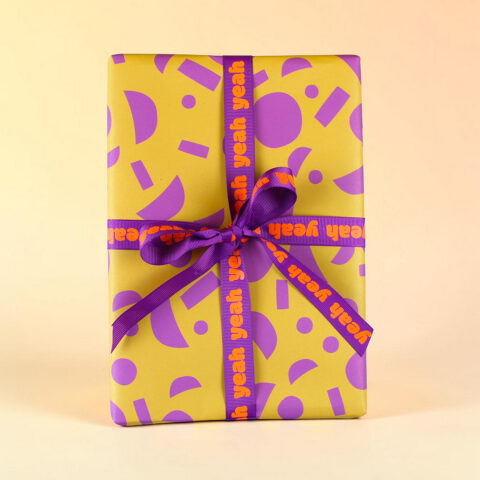 Yellow gift box with purple geometric wrap and a purple ribbon that reads 'yeah yeah yeah' in orange letters on the ribbon.