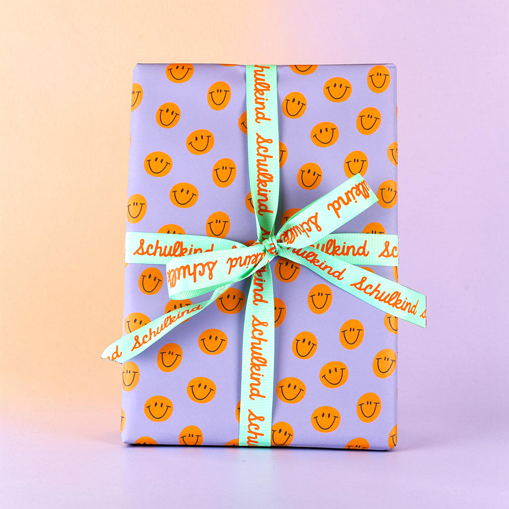Gift box wrapped in lavender paper with orange smiley faces, tied with a turquoise ribbon bow that reads 'Schulkind'.