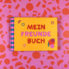 Bright yellow book titled 'MEIN FREUNDEBUCH' with purple and red text, blue-pink striped spine, on a pink patterned background with orange shapes.