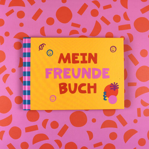 Bright yellow book titled 'MEIN FREUNDEBUCH' with purple and red text, blue-pink striped spine, on a pink patterned background with orange shapes.