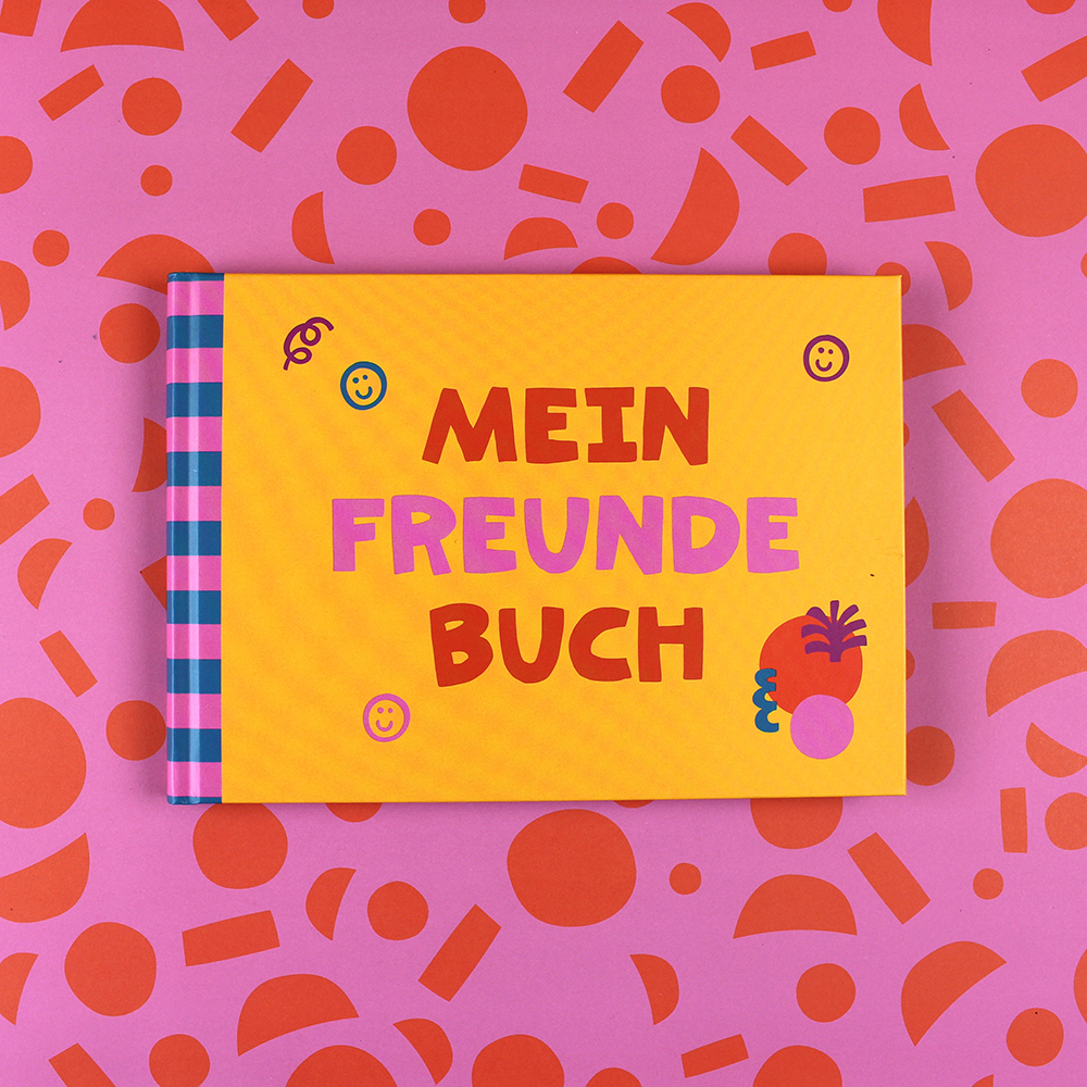 Bright yellow book titled 'MEIN FREUNDEBUCH' with purple and red text, blue-pink striped spine, on a pink patterned background with orange shapes.
