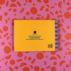 Yellow rectangular package on a pink patterned background with a small black logo and German text about recycled paper and plant-based inks.