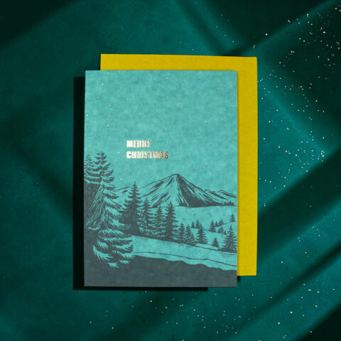 Turquoise Christmas card with a mountain and pine tree illustration and the words 'Merry Christmas' in white, layered over a yellow backing on a teal speckled background.
