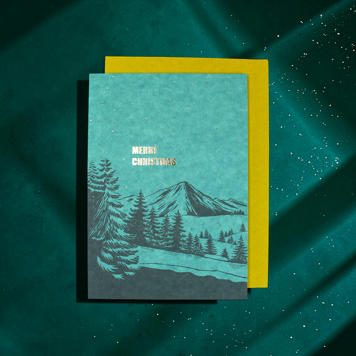 Turquoise Christmas card with a mountain and pine tree illustration and the words 'Merry Christmas' in white, layered over a yellow backing on a teal speckled background.