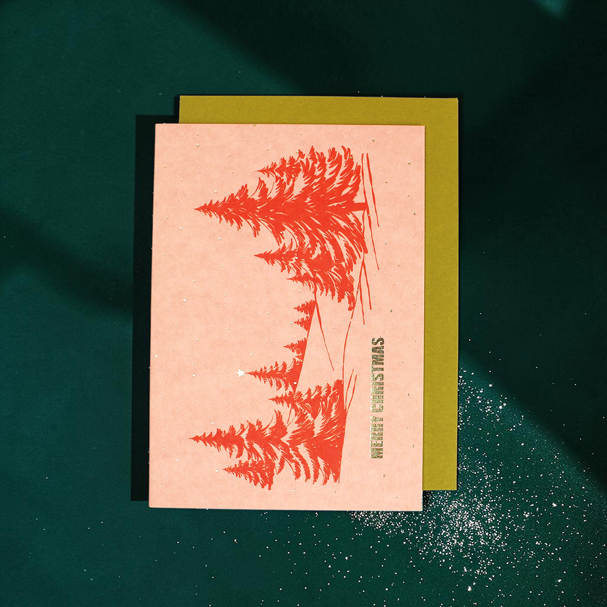 2364grusskartemerrychristmasmountainsrosa Avayves Feine Papeterie Und Geschenke Für Babys Und Kids Pink Christmas card with red fern graphic, stacked on an olive card, on a dark green surface with white speckles and glitter hints.
