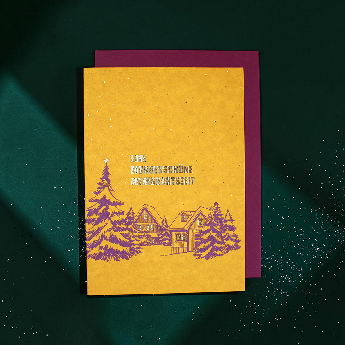 Yellow holiday card with purple winter village illustration on a green background; German text reads 'Eine wunder schöne Weihnachtszeit' and a purple backing shows behind.