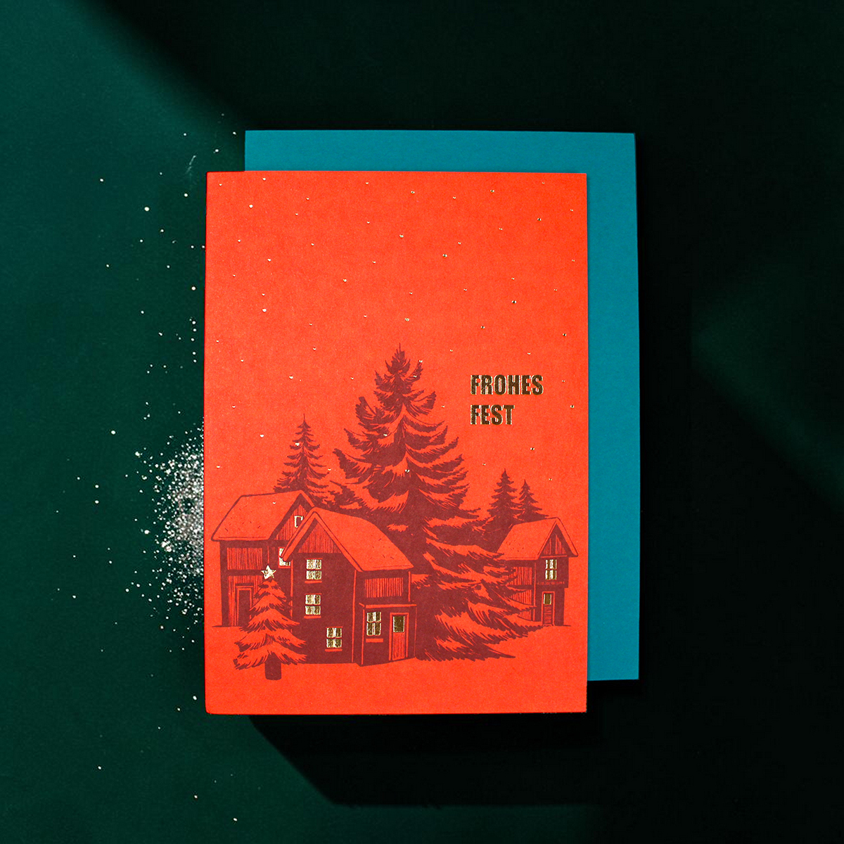 Red holiday card with a winter scene of houses and evergreen trees; says 'FROHES FEST' in bold letters, blue card behind.