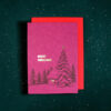 Pink Christmas card with gold 'Merry Christmas' text, a small cabin and pine trees in a snowy forest scene, set on a dark green speckled background with a red envelope beneath.