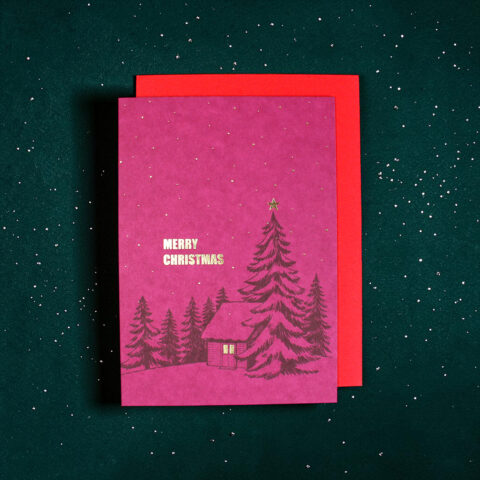 Pink Christmas card with gold 'Merry Christmas' text, a small cabin and pine trees in a snowy forest scene, set on a dark green speckled background with a red envelope beneath.