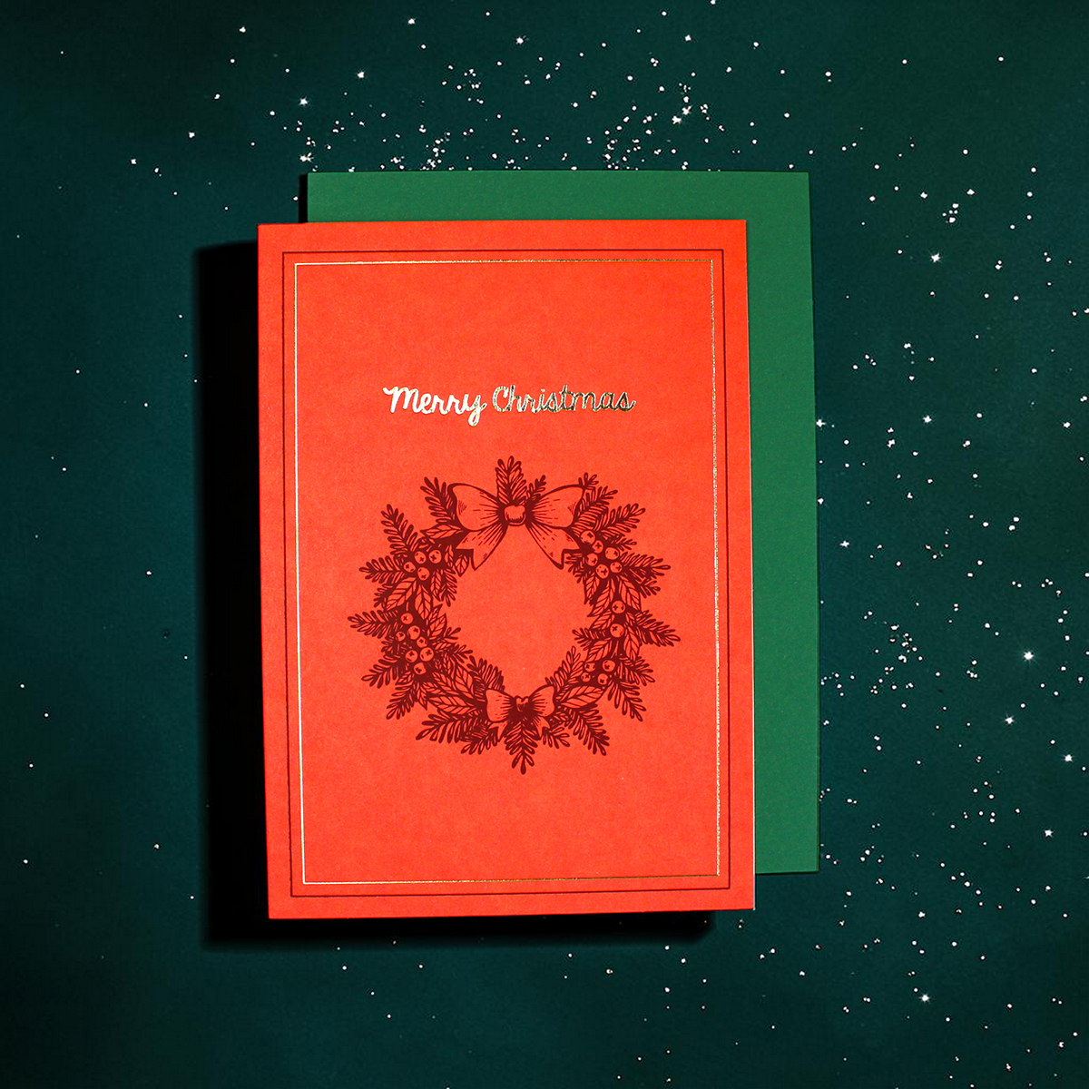 Red Christmas card with a wreath illustration and 'Merry Christmas' on the front, placed on a green envelope against a snow-speckled dark background.