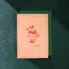 Pink Christmas card featuring a red duck wearing a knit hat and scarf, with 'Merry Christmas' text, placed on a green envelope on a dark green snowy background.