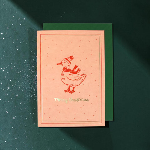 Pink Christmas card featuring a red duck wearing a knit hat and scarf, with 'Merry Christmas' text, placed on a green envelope on a dark green snowy background.