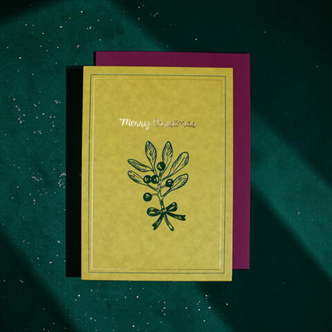 Yellow Christmas card with a leafy botanical illustration and 'Merry Christmas' text on a teal surface with a magenta backing.