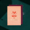 2372grusskarteweihnachtenpilzanhaenger Avayves Feine Papeterie Und Geschenke Für Babys Und Kids Peach Christmas card with a red mushroom illustration tied with a bow and gold 'Merry Christmas' text, layered on a burgundy card on a green background.