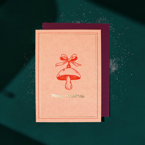 Peach Christmas card with a red mushroom illustration tied with a bow and gold 'Merry Christmas' text, layered on a burgundy card on a green background.