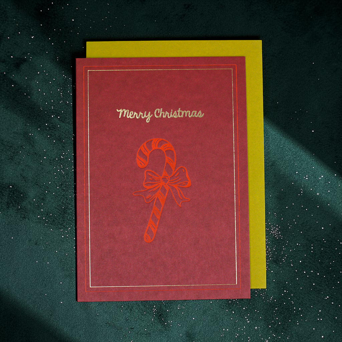 Red Christmas card with gold 'Merry Christmas' text and a candy cane with a bow on a green speckled background, backed by yellow card.
