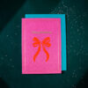 Pink Christmas card with a red bow illustration and 'Merry Christmas' in gold, resting on a turquoise backing with scattered white confetti.