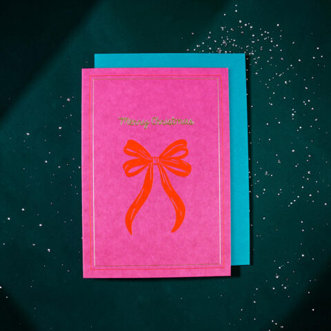 Pink Christmas card with a red bow illustration and 'Merry Christmas' in gold, resting on a turquoise backing with scattered white confetti.