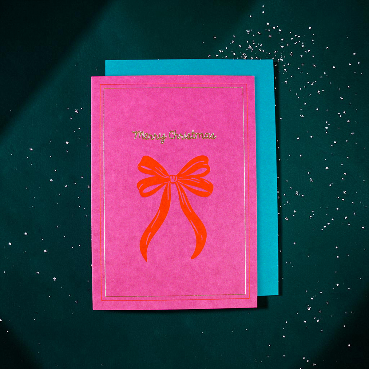 Pink Christmas card with a red bow illustration and 'Merry Christmas' in gold, resting on a turquoise backing with scattered white confetti.