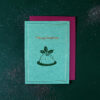 Teal Christmas card with holly motif and 'Merry Christmas' text, resting on a magenta envelope on a glittery dark green background.