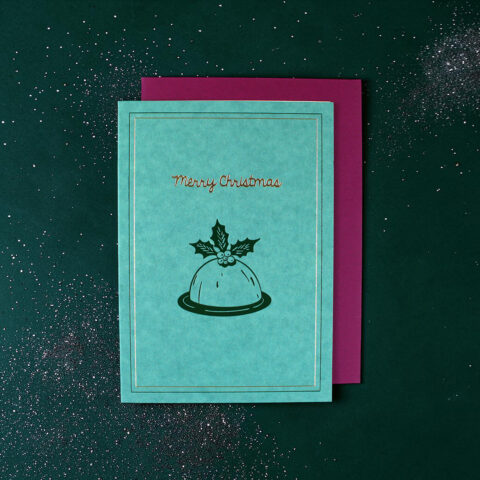 Teal Christmas card with holly motif and 'Merry Christmas' text, resting on a magenta envelope on a glittery dark green background.