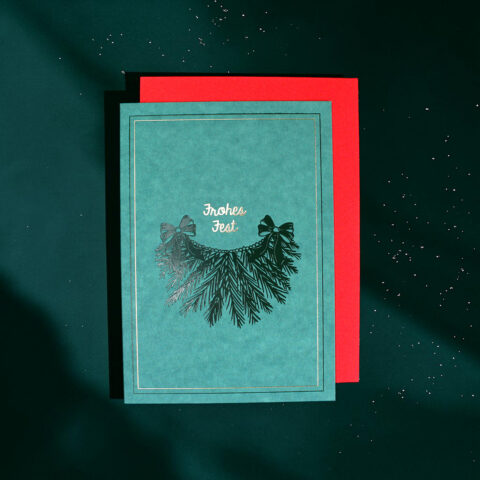 Teal Christmas card with gold text 'Frohes Fest' and a pine bow design on a red backing set against a dark green background.