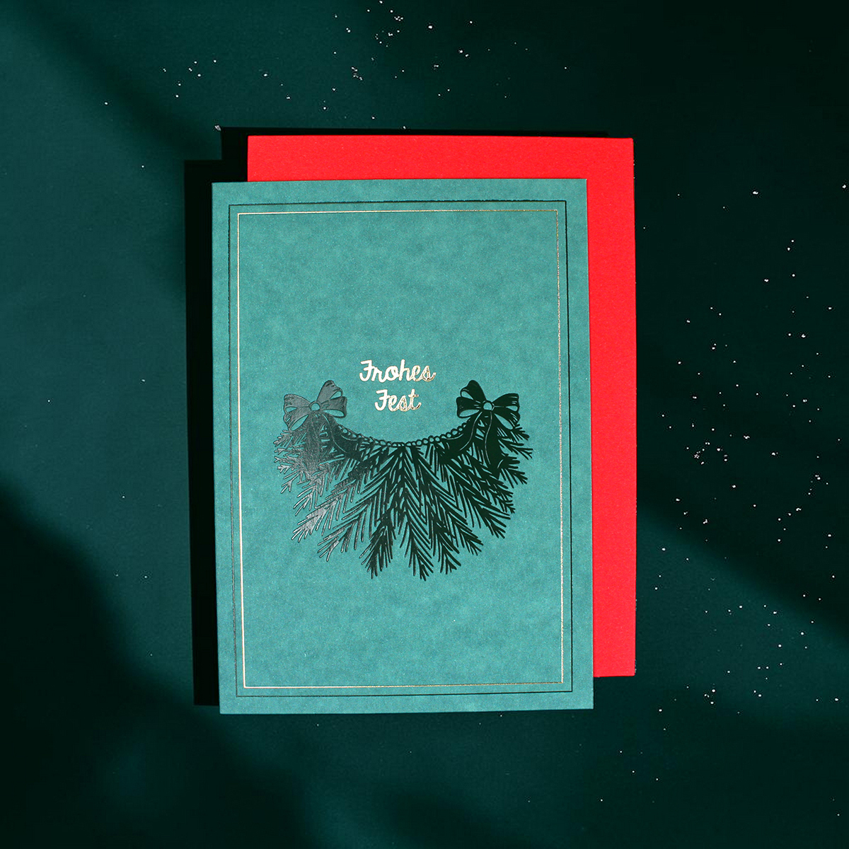 Teal Christmas card with gold text 'Frohes Fest' and a pine bow design on a red backing set against a dark green background.
