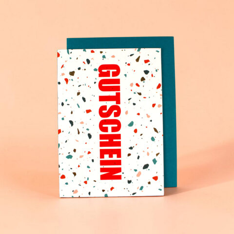 Terrazzo-patterned white gift card with bold red vertical word 'GUTSCHEIN' leaning against a teal envelope on a peach background, indicating a voucher.