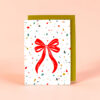 2378grusskarteb6schleifehell Avayves Feine Papeterie Und Geschenke Für Babys Und Kids White gift card with a large red bow and colorful confetti pattern, propped in front of a green envelope on a peach background (greeting card).