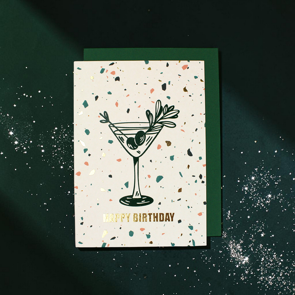 2380grusskarteb6happybirthday Avayves Feine Papeterie Und Geschenke Für Babys Und Kids Birthday card with a cocktail glass illustration and confetti on a white card over a green envelope on a dark background, reads 'Happy Birthday' in gold letters.