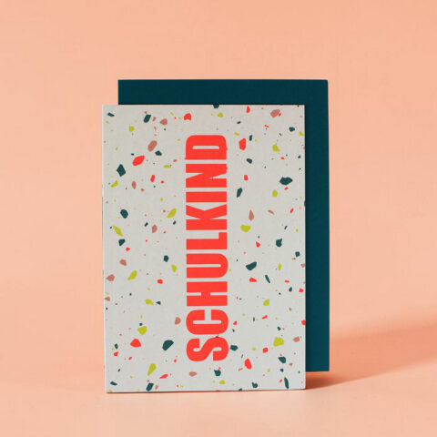 Greeting card with a speckled confetti pattern and bold vertical red text 'SCHULKIND' leaning against a dark teal panel on a peach background.