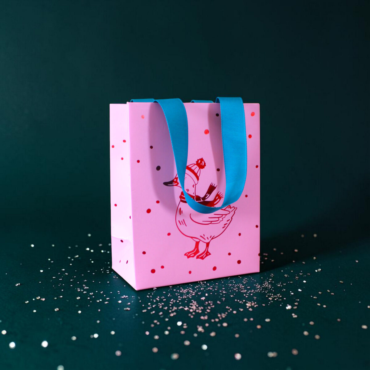 4176geschenktaschesgans Avayves Feine Papeterie Und Geschenke Für Babys Und Kids Pink gift bag with bright blue handles resting on a dark teal surface, dotted with small white confetti pieces.