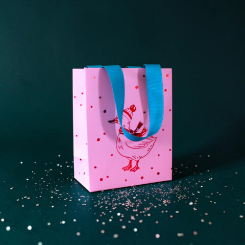 Pink gift bag with bright blue handles resting on a dark teal surface, dotted with small white confetti pieces.