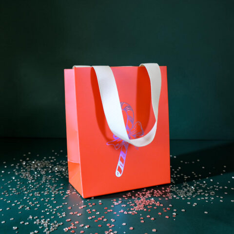 Red gift bag with white handles set against a dark green background, with scattered red and white confetti around it.