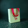 4178geschenktaschesmistelzweig Avayves Feine Papeterie Und Geschenke Für Babys Und Kids Pale green gift bag with bright red handles on a dark green textured background, sprinkled with white confetti.