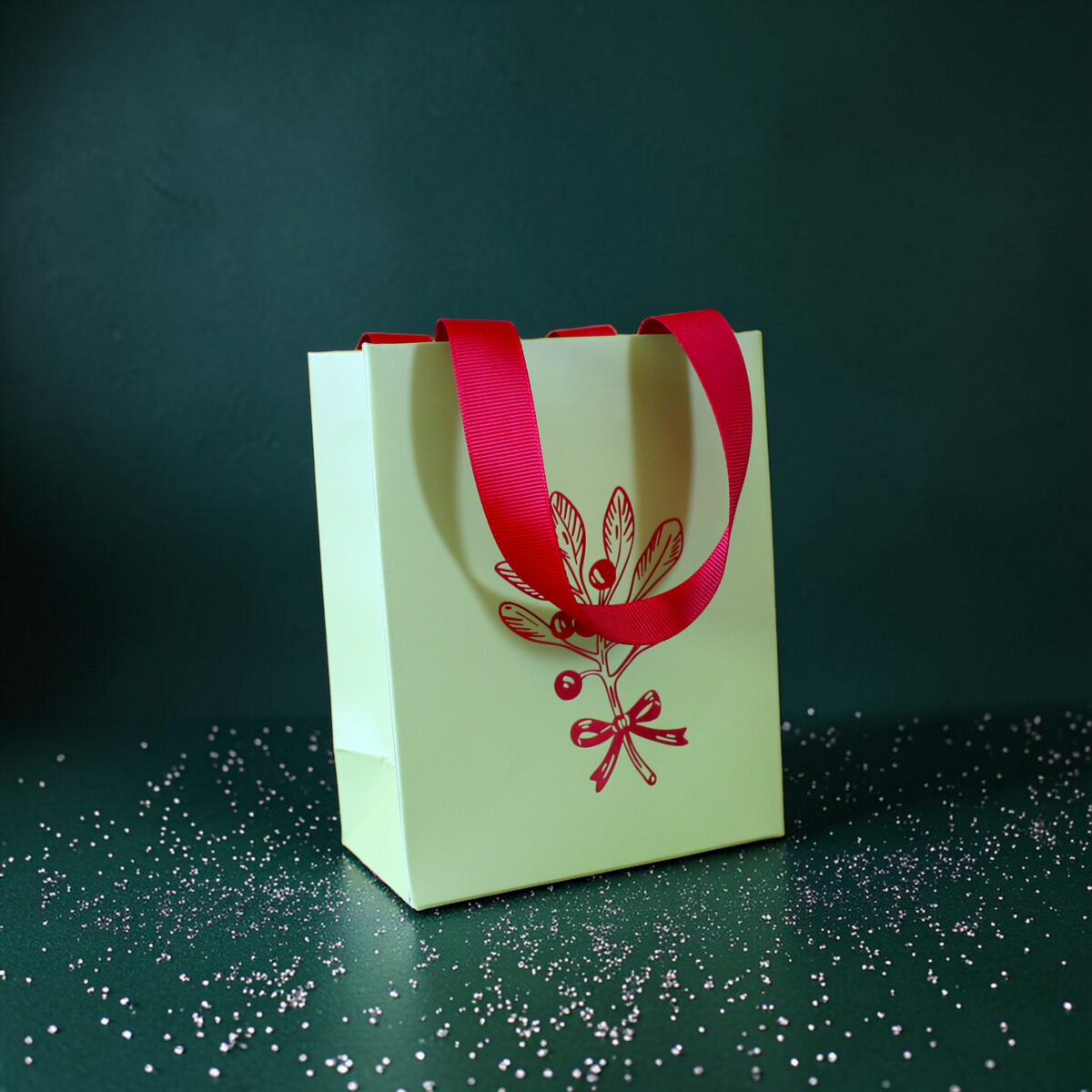 4178geschenktaschesmistelzweig Avayves Feine Papeterie Und Geschenke Für Babys Und Kids Pale green gift bag with bright red handles on a dark green textured background, sprinkled with white confetti.