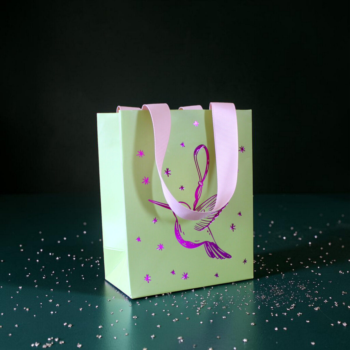 4179geschenktaschesvogel Avayves Feine Papeterie Und Geschenke Für Babys Und Kids Pastel green gift bag with pink handles and purple star decorations on a dark background with scattered confetti.